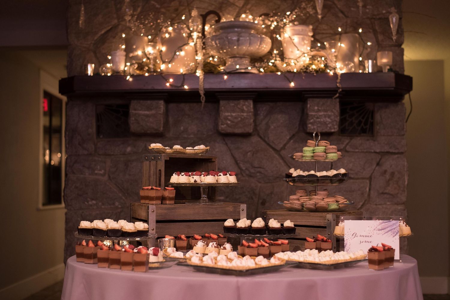 Elegant dessert table with tiered treats displayed before a stone fireplace adorned with warm string lights.