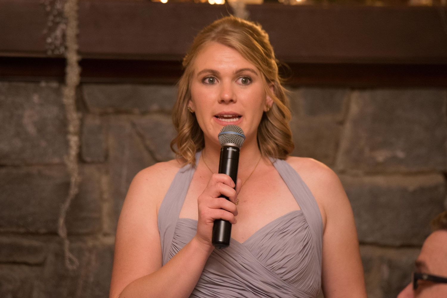 Woman in gray bridesmaid dress giving a speech at a wedding reception while holding a microphone.