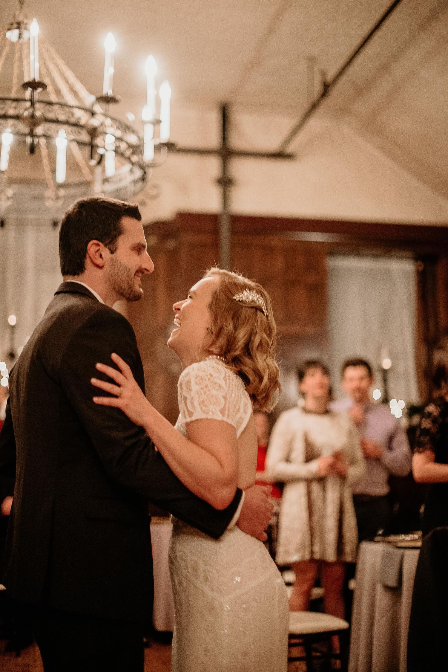 A bride and groom share their first dance, smiling joyfully, beneath a chandelier at their wedding reception.