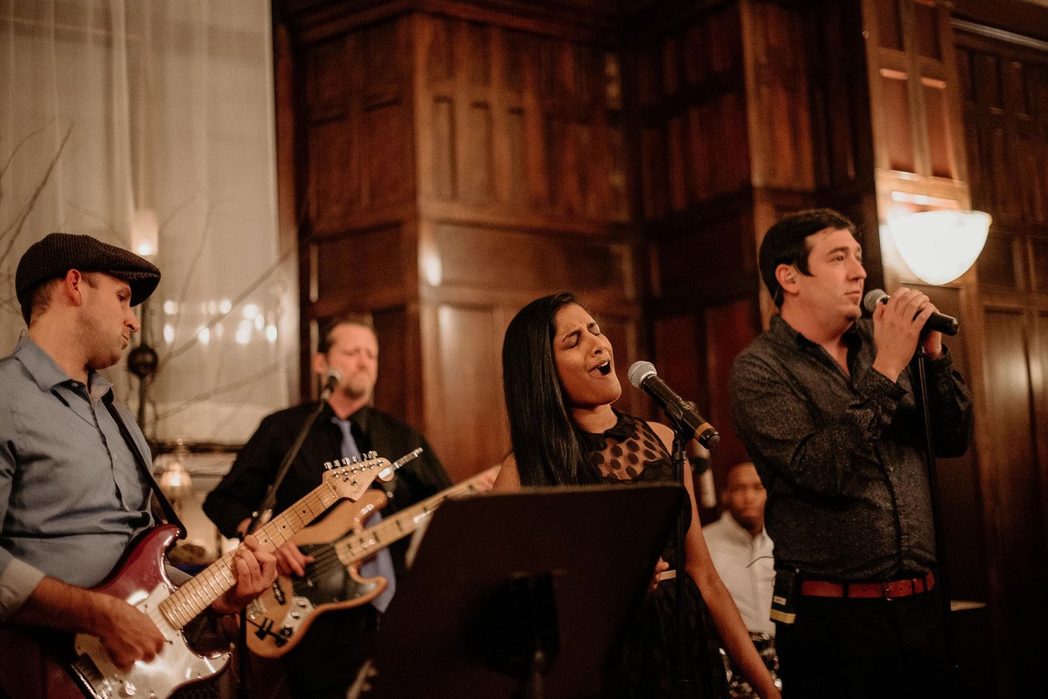 Live band performing at elegant wood-paneled venue, featuring guitarist, female vocalist, and male singer with microphone.