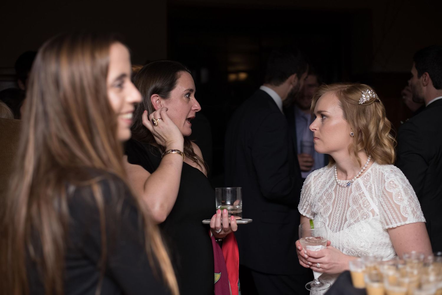 Women in formal attire socializing at an elegant evening event, holding wine glasses at what appears to be a wedding reception.