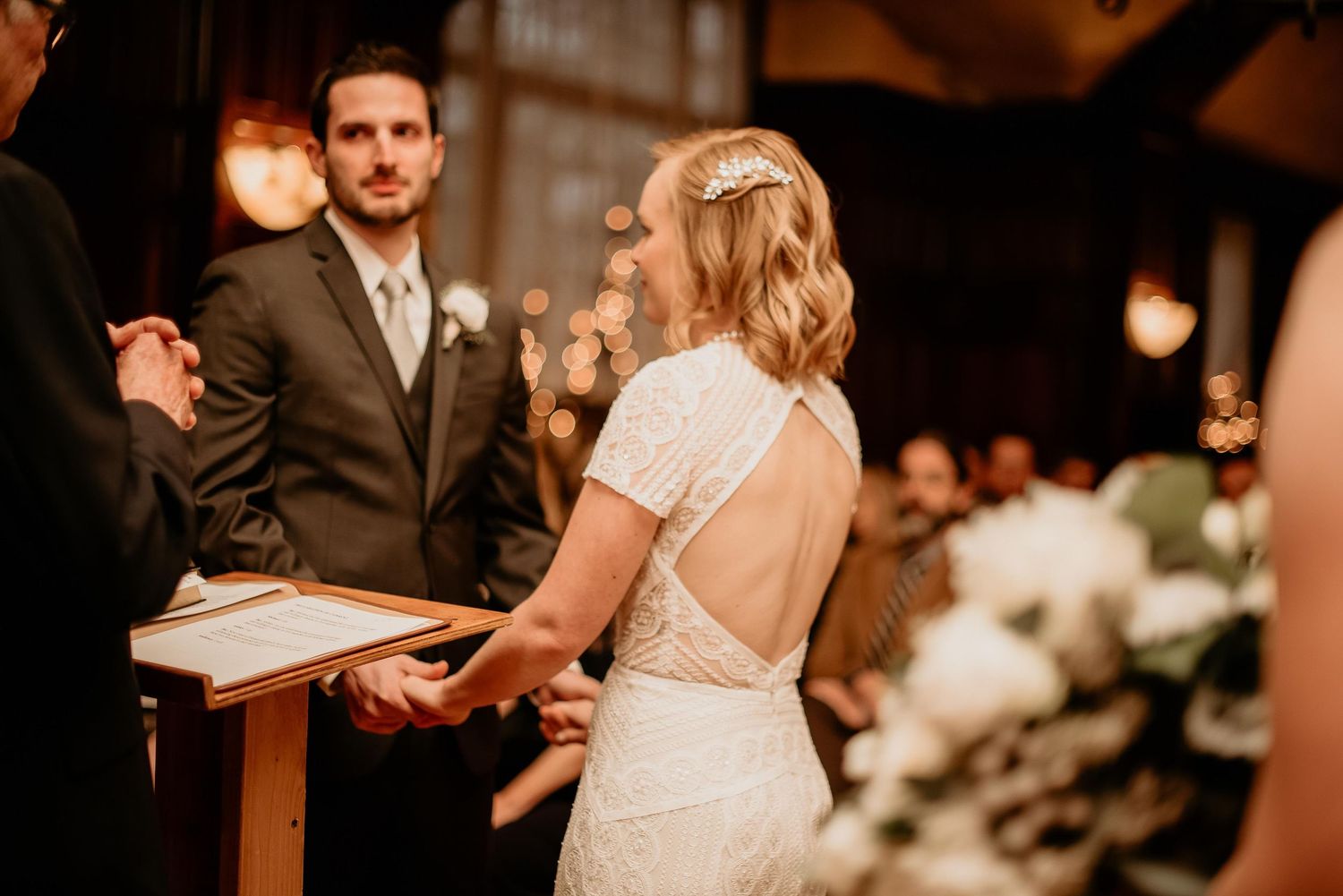 Bride in lace backless wedding dress holds hands with groom during indoor wedding ceremony.