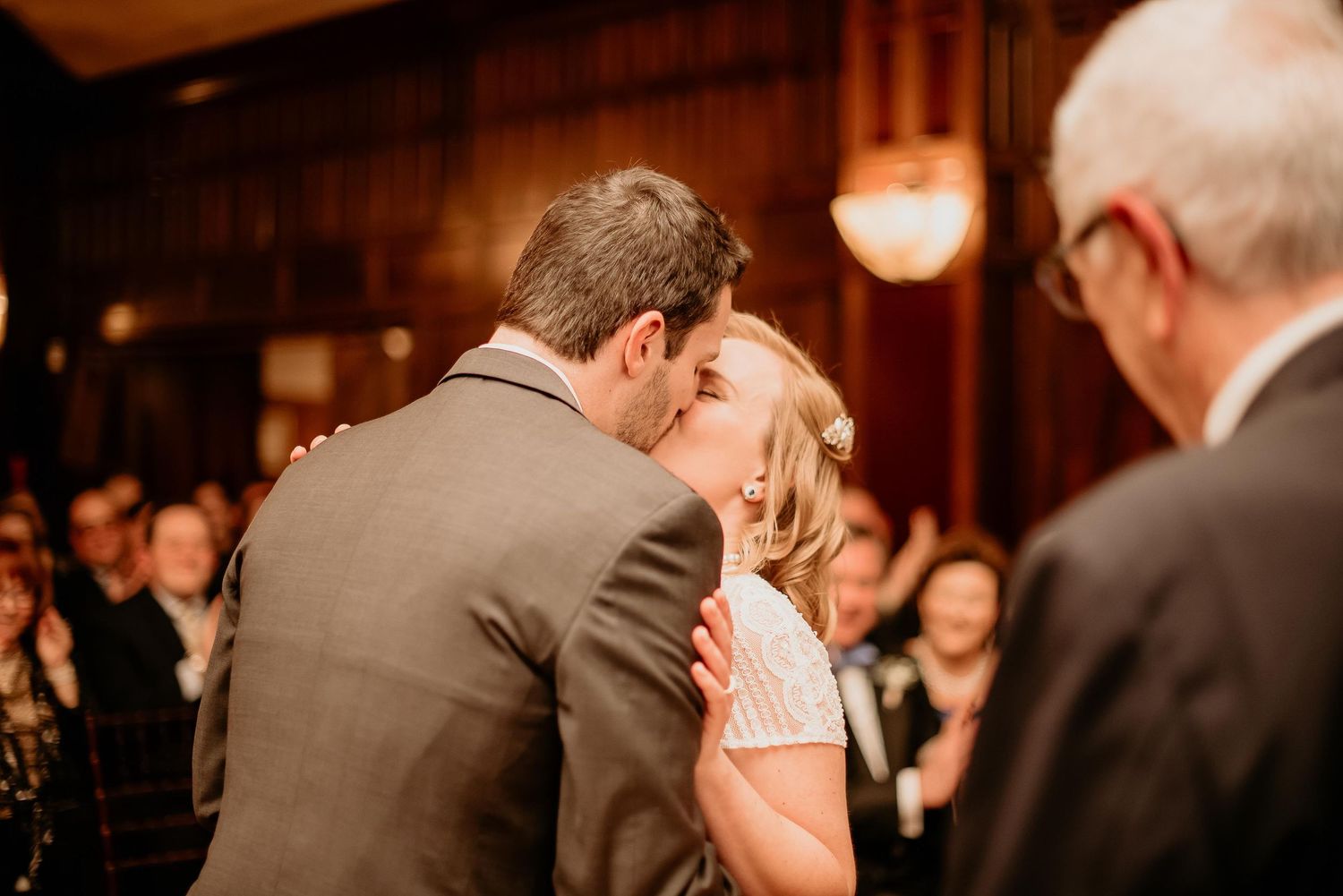 Bride and groom share their first kiss at wedding ceremony with guests applauding in elegant wood-paneled venue.