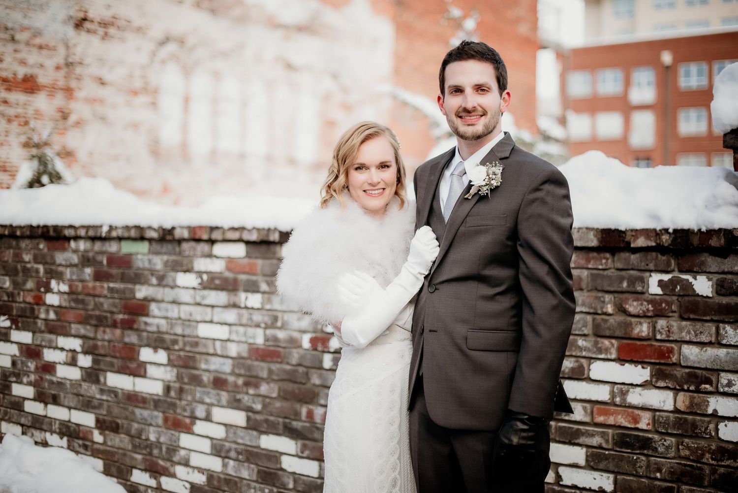 Bride and groom posing together in winter snow beside a brick wall, bride in white fur wrap, groom in gray suit.