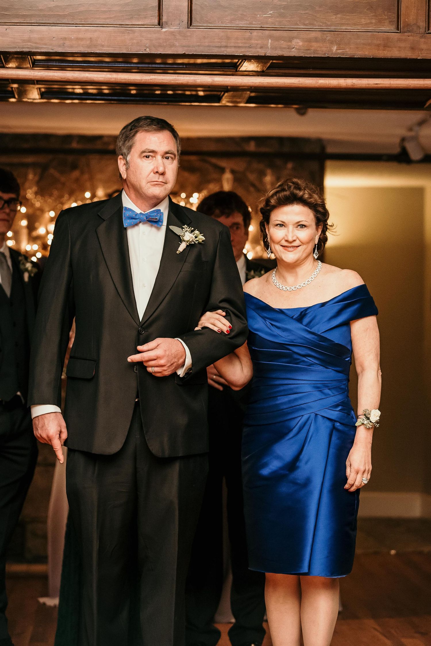 Elegant couple in formal attire, man in black tuxedo with blue bow tie, woman in royal blue off-shoulder dress, at wedding.