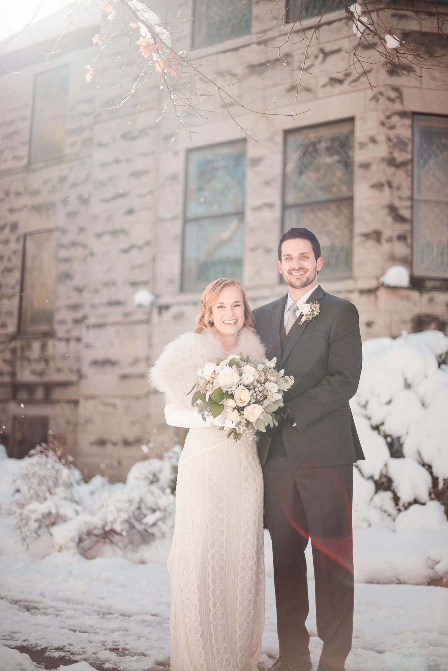 Bride and groom posing in snow outside stone church, bride holding winter bouquet wearing fur wrap and knit gown.