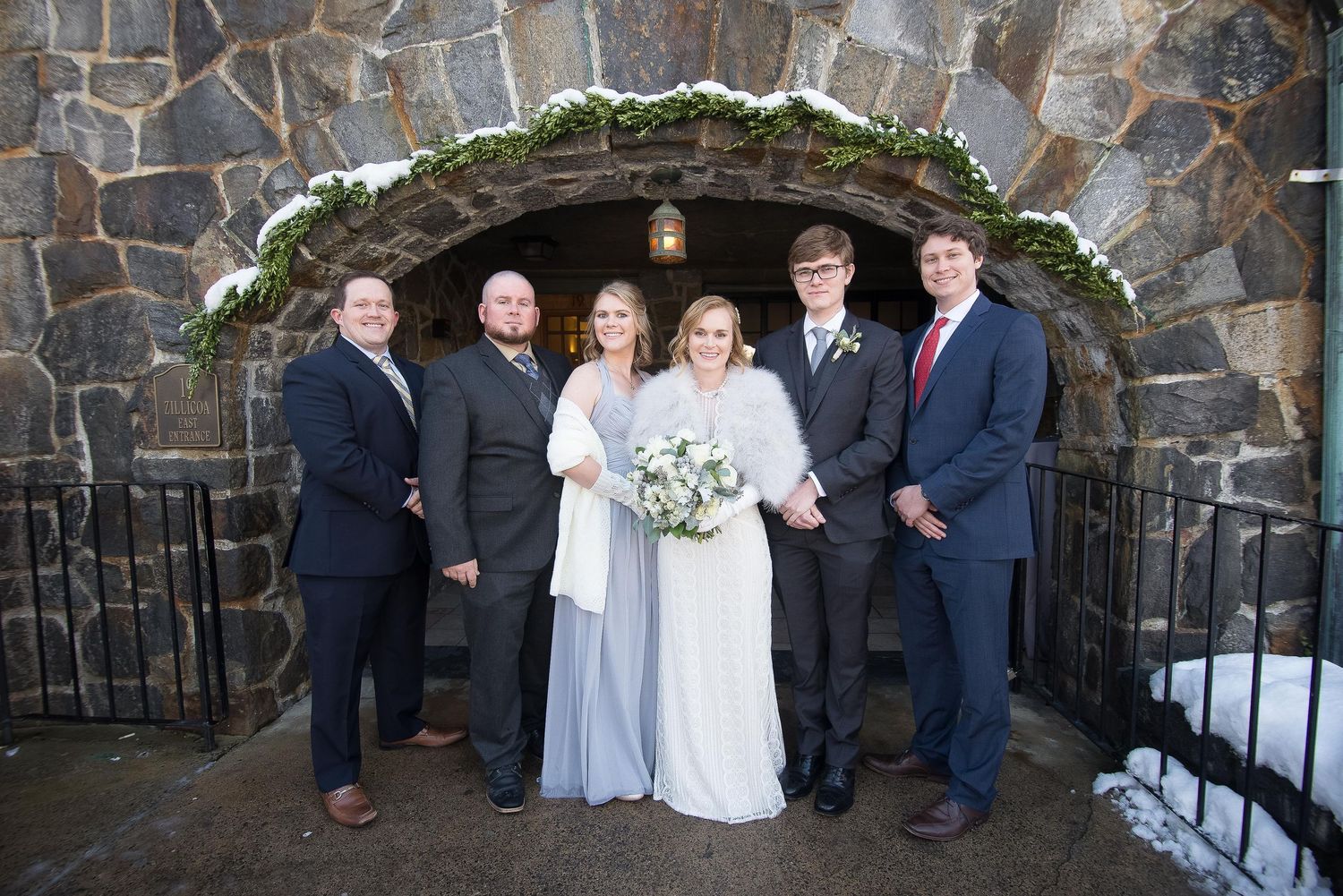 Wedding party posing in front of a snow-dusted stone arch entrance in winter.