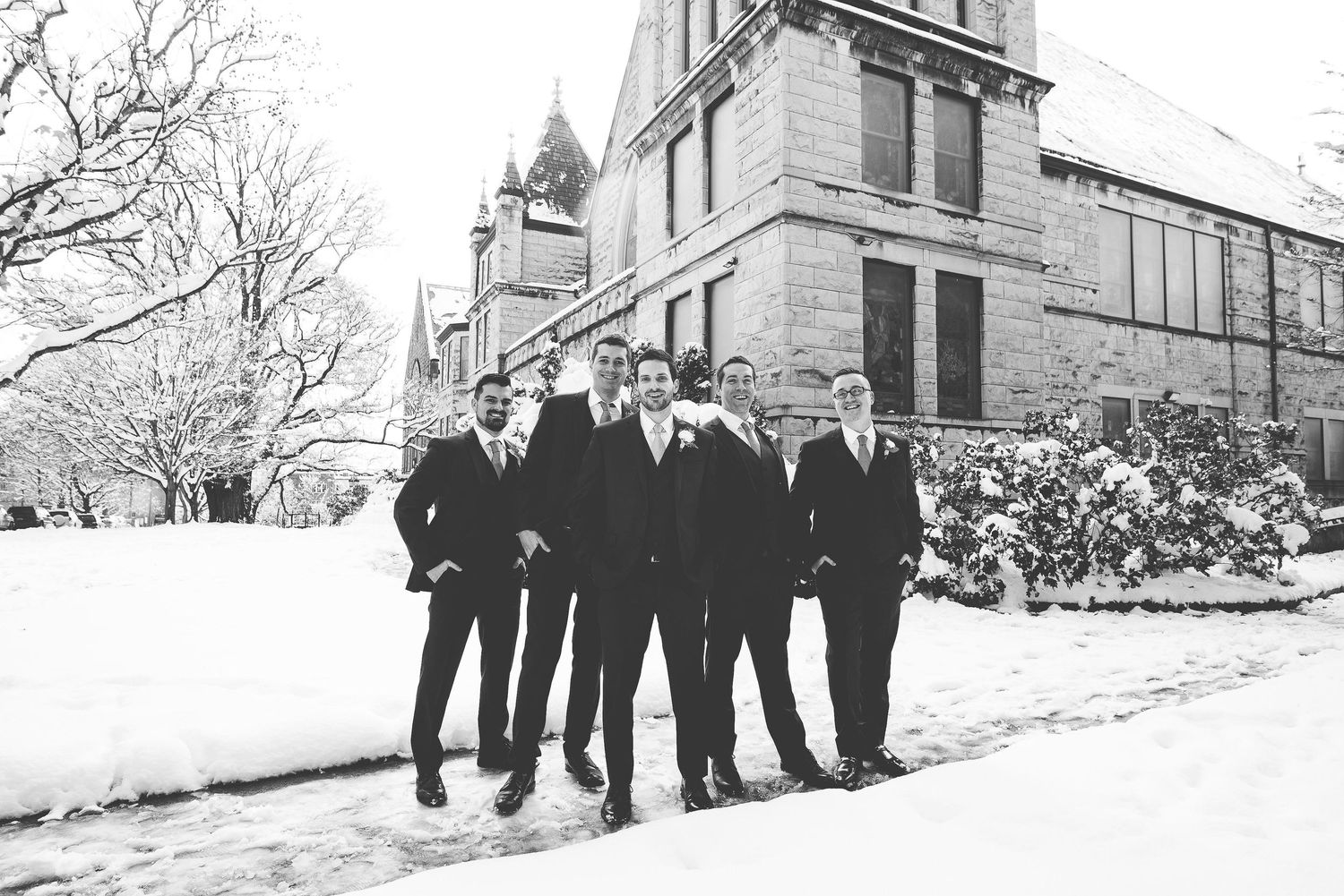 Five groomsmen in black suits pose together in snowy winter wedding photo outside historic stone building.