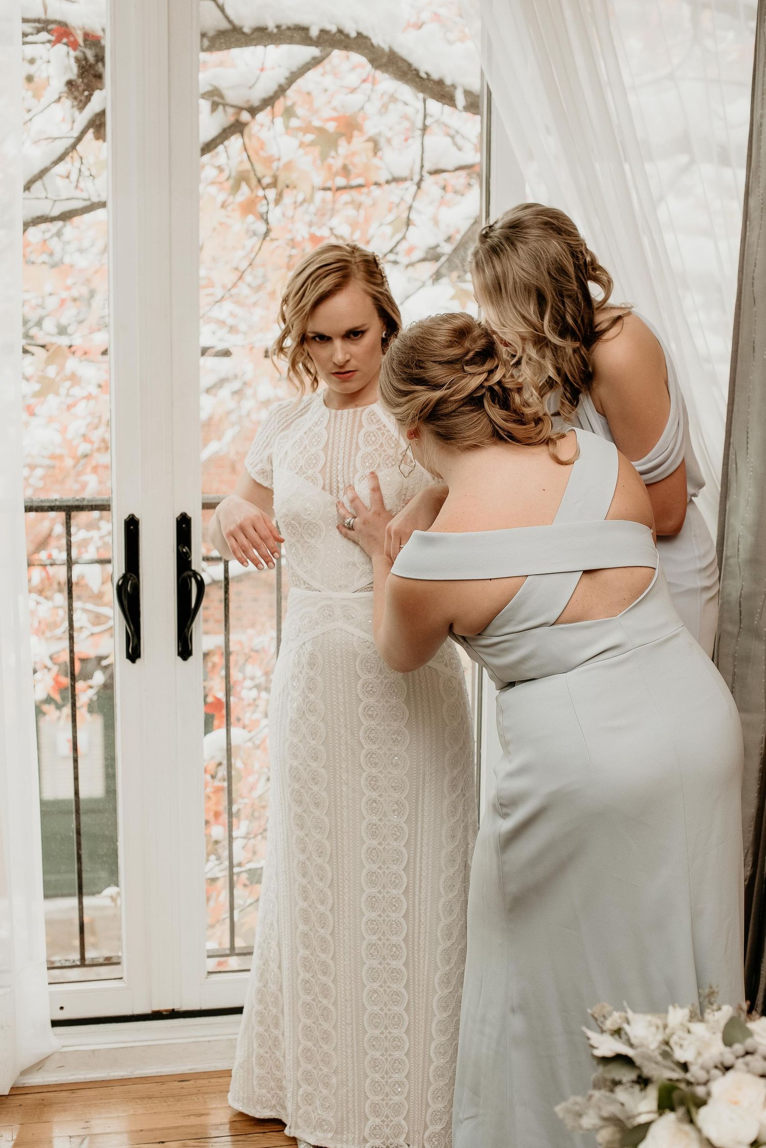 Bride in lace gown being assisted by two bridesmaids in sage dresses near French doors with autumn foliage.