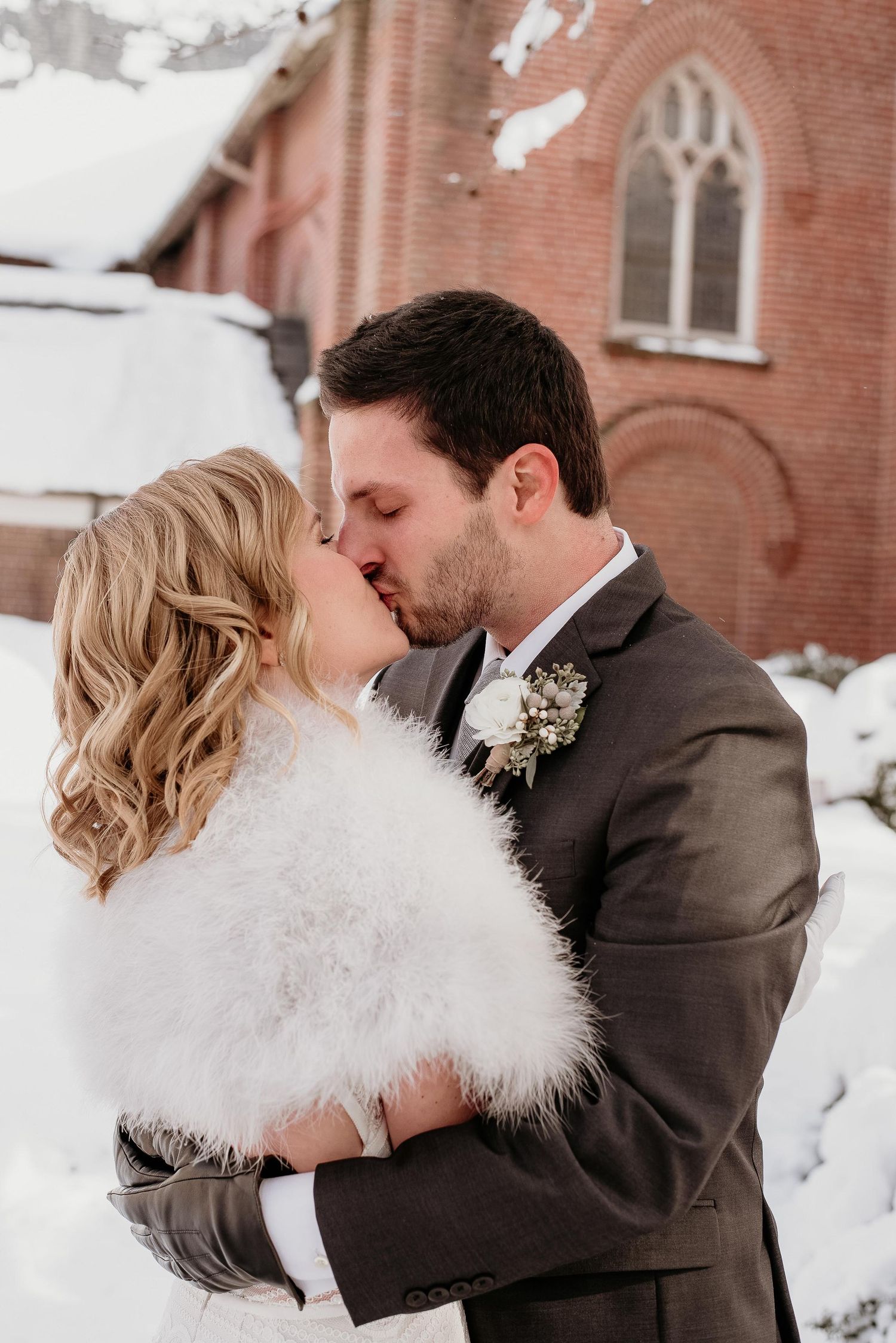 Bride and groom share a romantic kiss in the snow outside a brick church, bride wearing a white fur wrap.