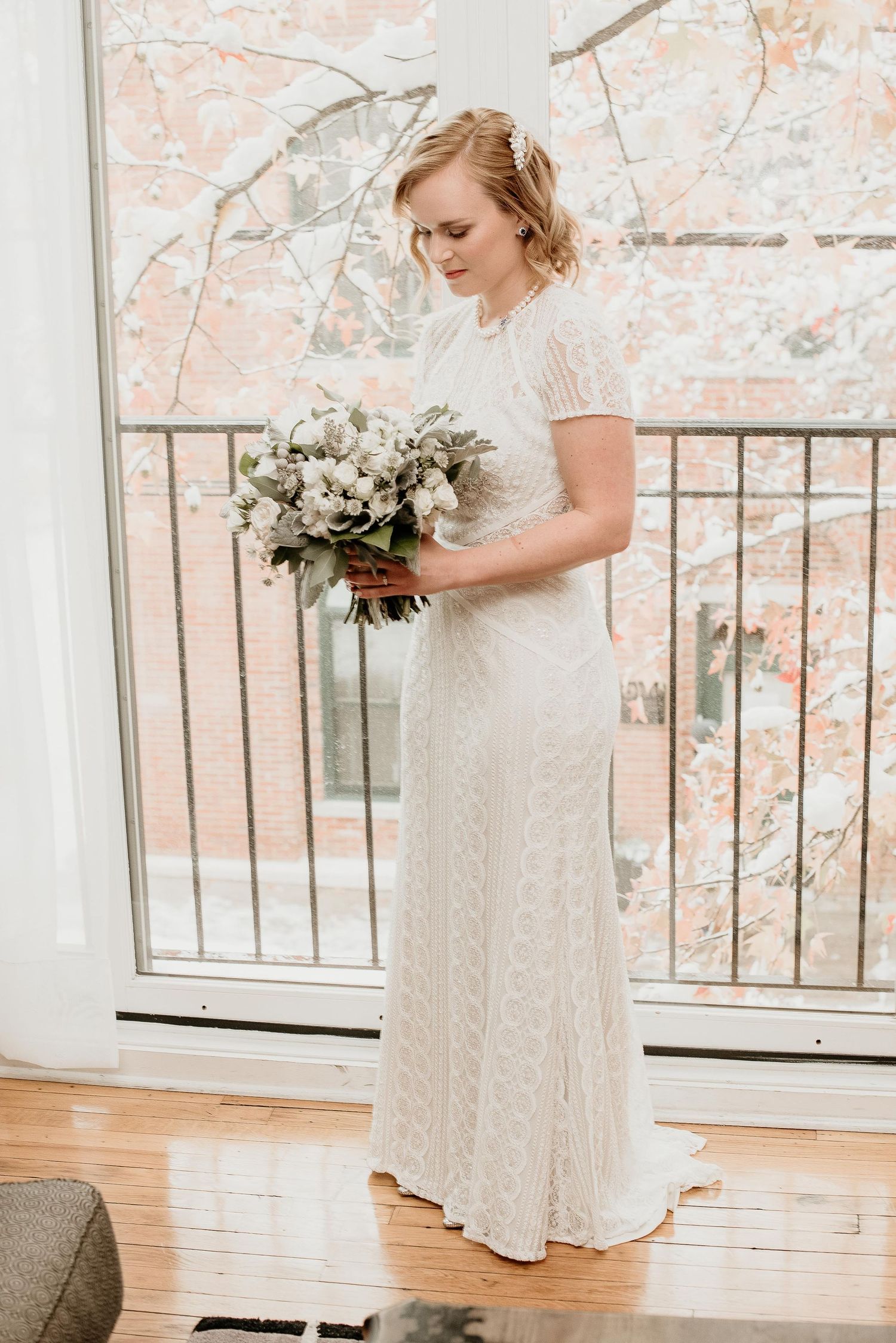 Bride in white lace wedding gown holding bouquet of white flowers, standing by large window with winter trees.