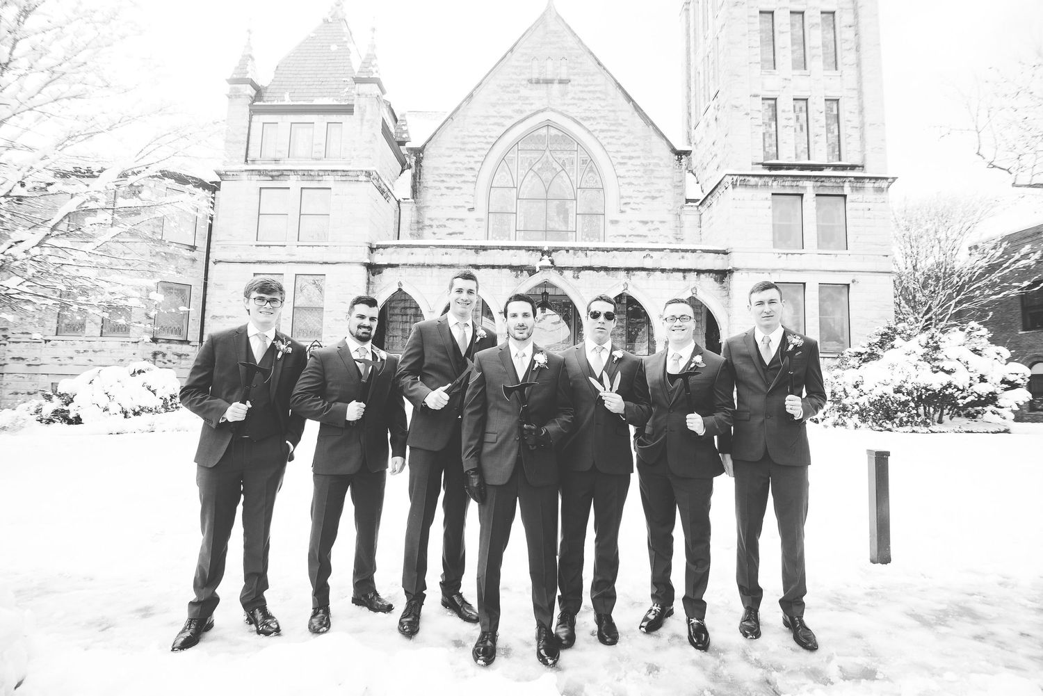 Groomsmen in dark suits posing in snow outside a historic stone church building in a black and white wedding photo.