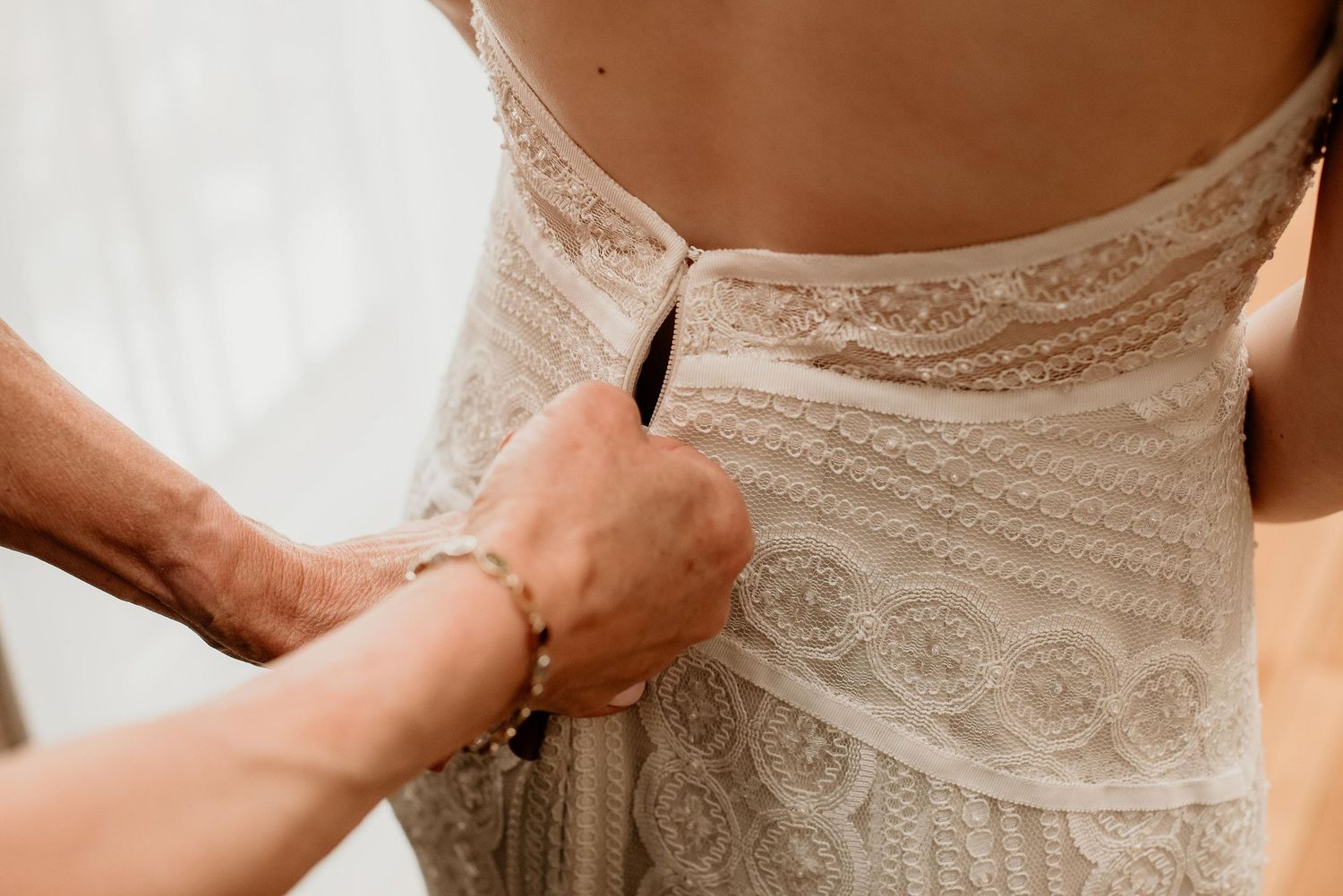 A bride getting her lace wedding dress buttoned up from behind by a helping hand wearing a gold bracelet.
