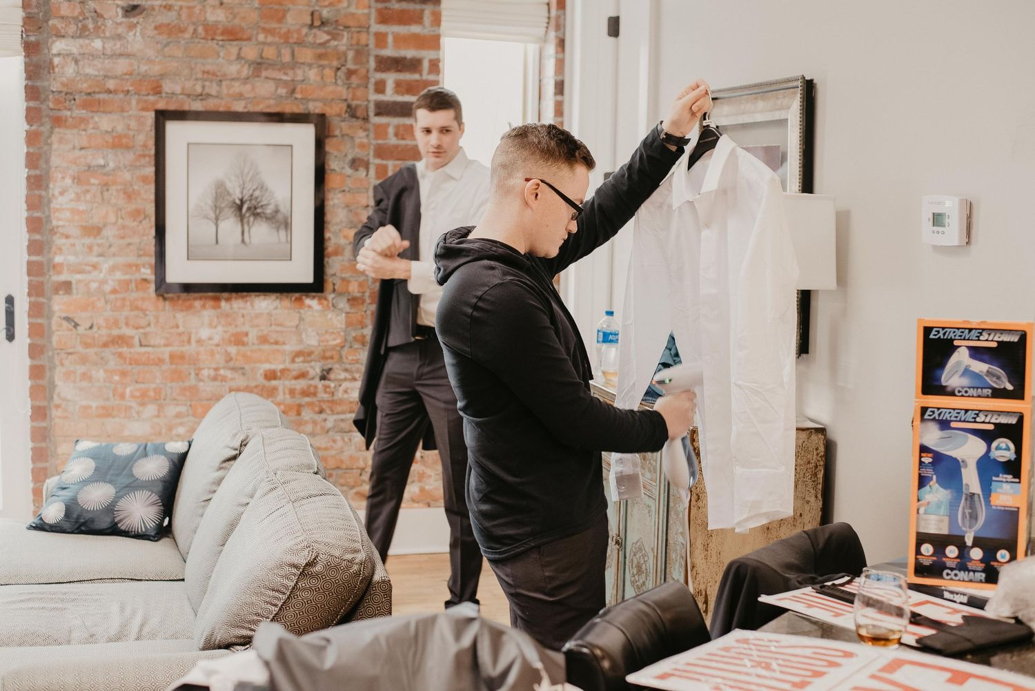 Two men in a modern apartment with brick walls preparing formal wear, one holding up a dry-cleaned shirt in plastic.