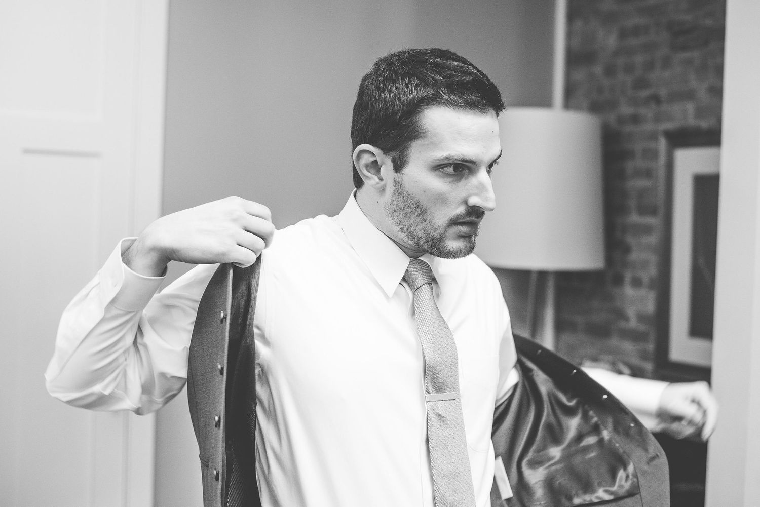 Black and white photo of a well-dressed man putting on a suit jacket while wearing a white shirt and tie.