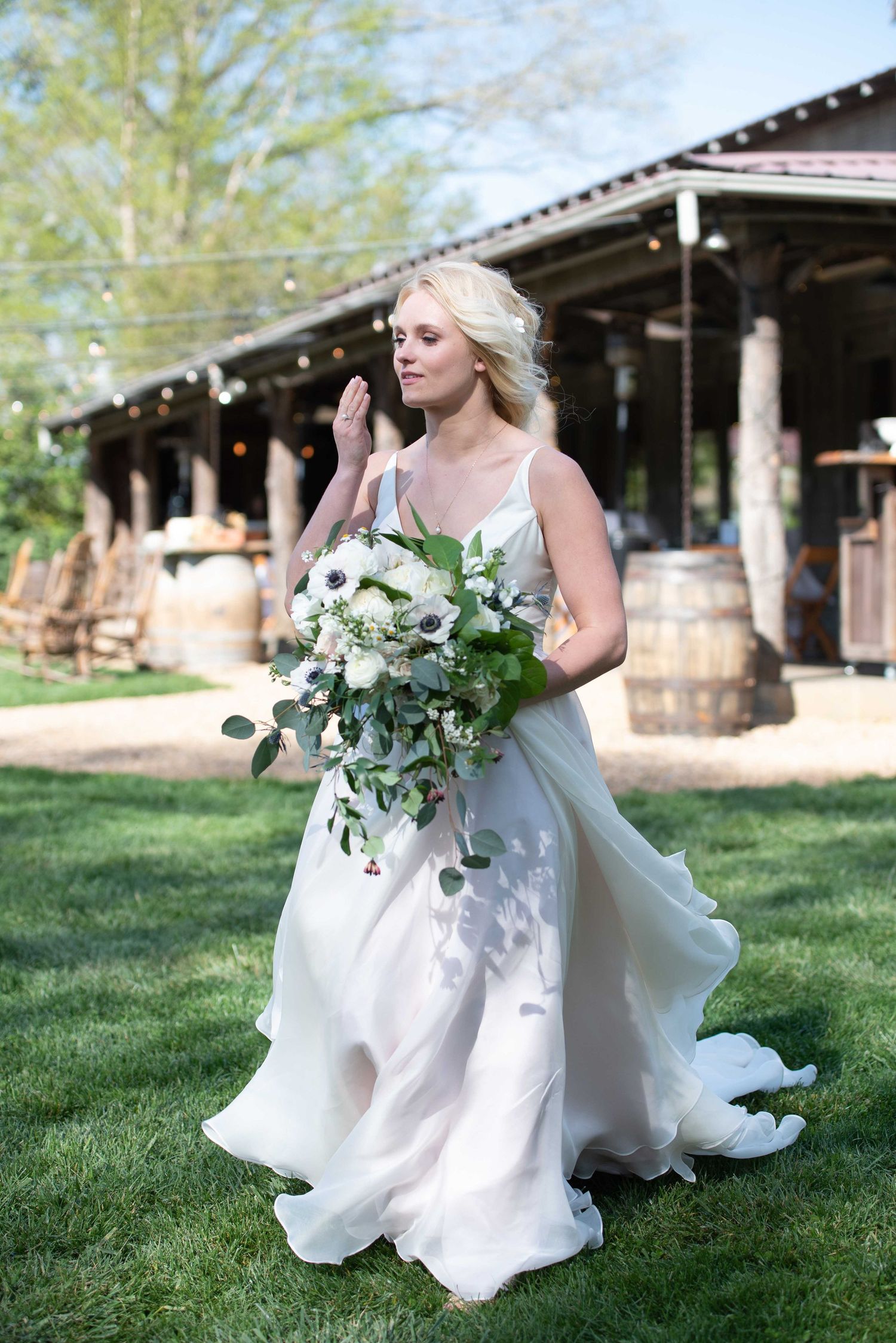 Bride in flowing white gown holds green and white bouquet outdoors near rustic barn venue.