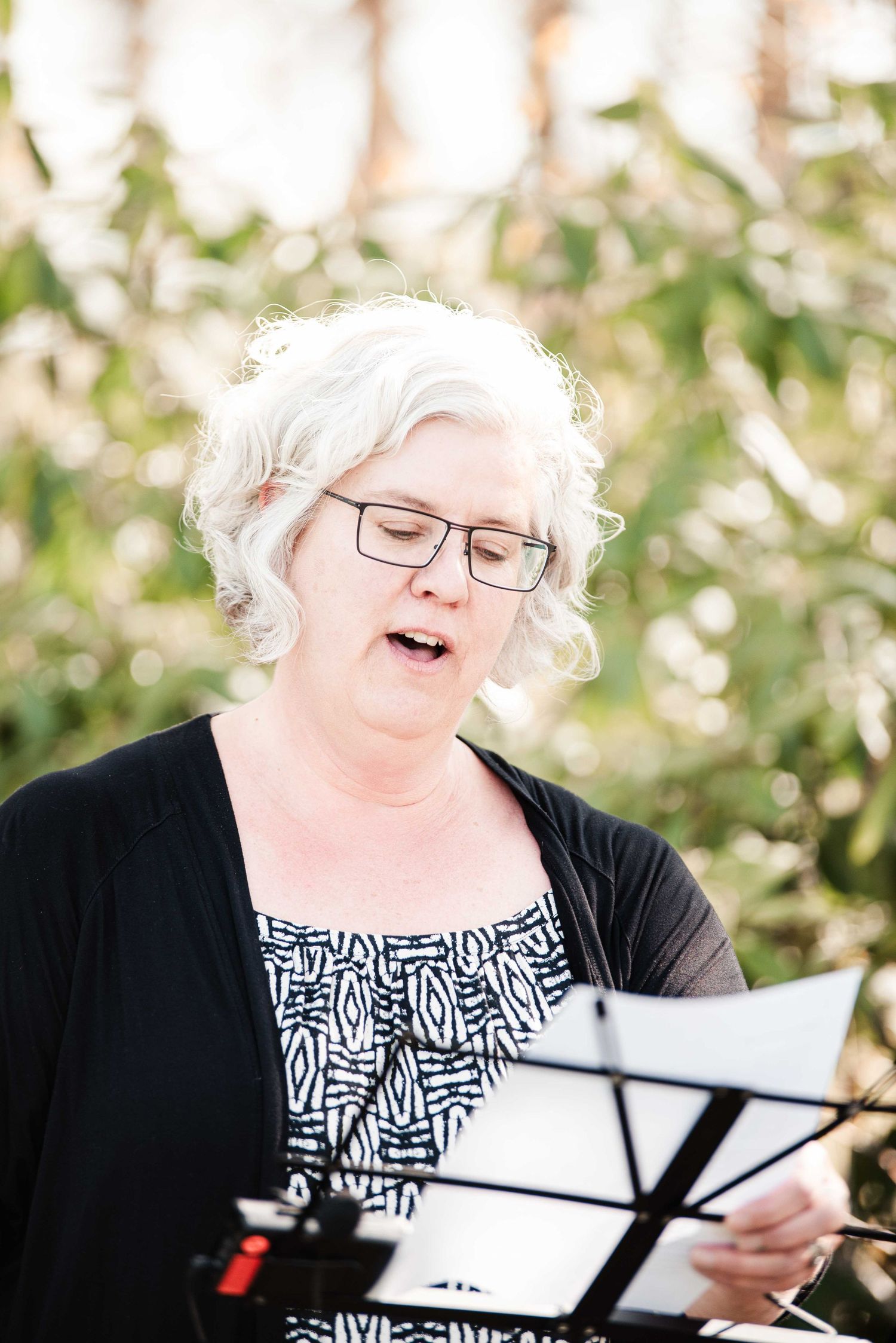 Older woman with white hair and glasses singing outdoors, holding sheet music at a music stand.