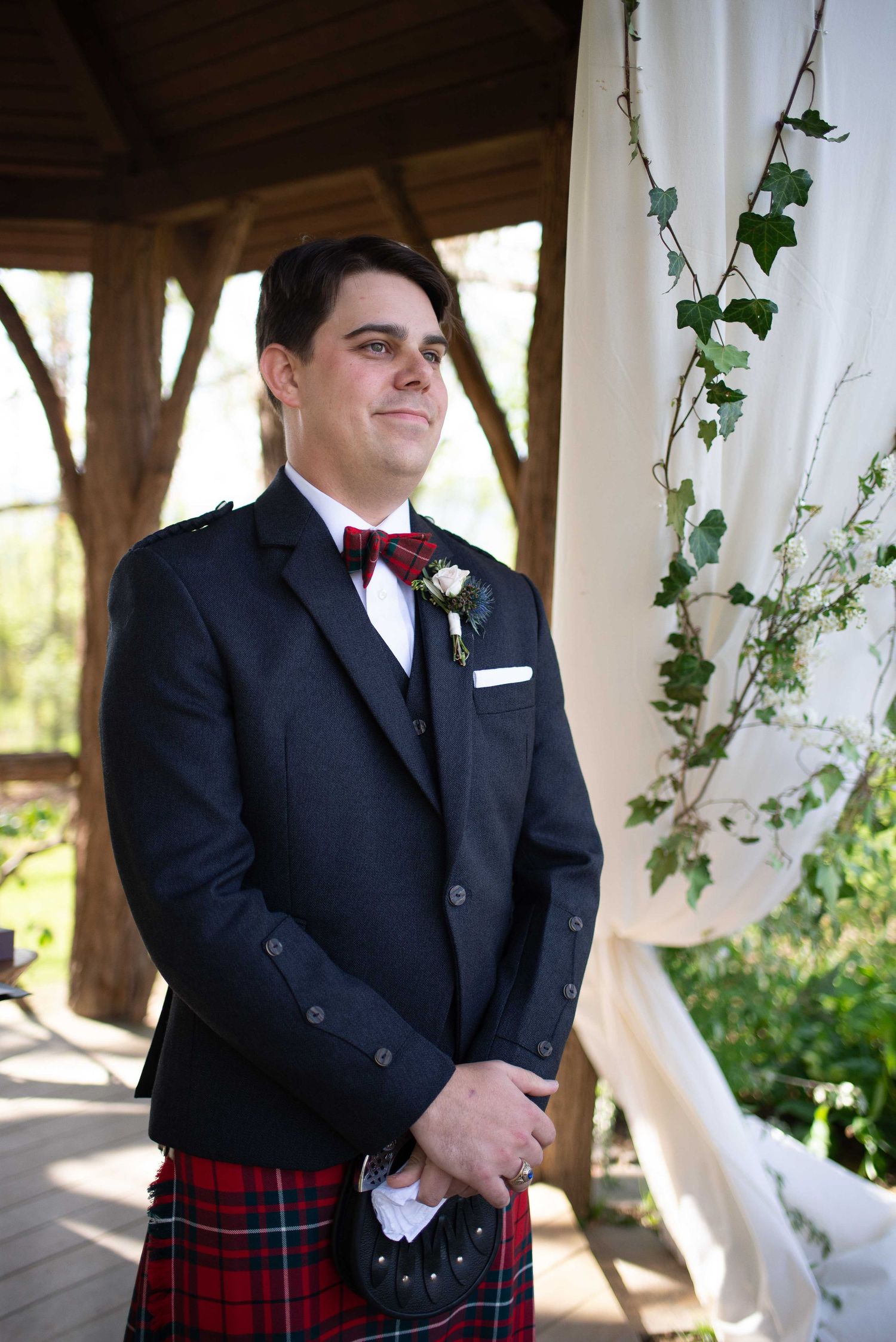 Groom in tartan kilt and dark jacket with boutonniere smiles at wedding ceremony altar with ivy decor.