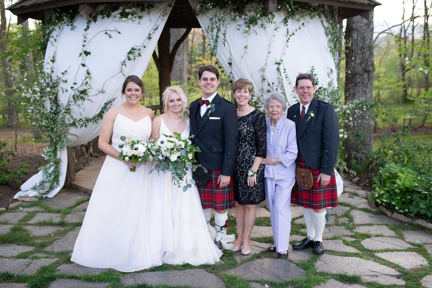 Wedding party posing under floral gazebo, bride in white gown, two men in red kilts, outdoors on stone path.