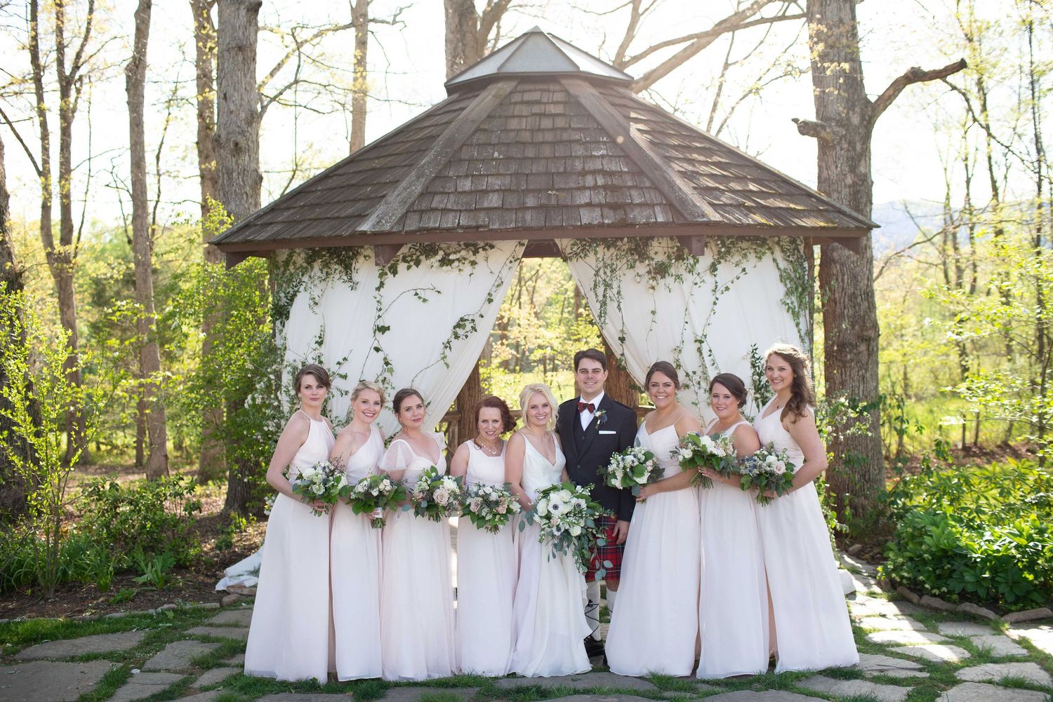 Wedding party poses under floral gazebo with bridesmaids in blush gowns and groom in kilt surrounded by lush greenery.