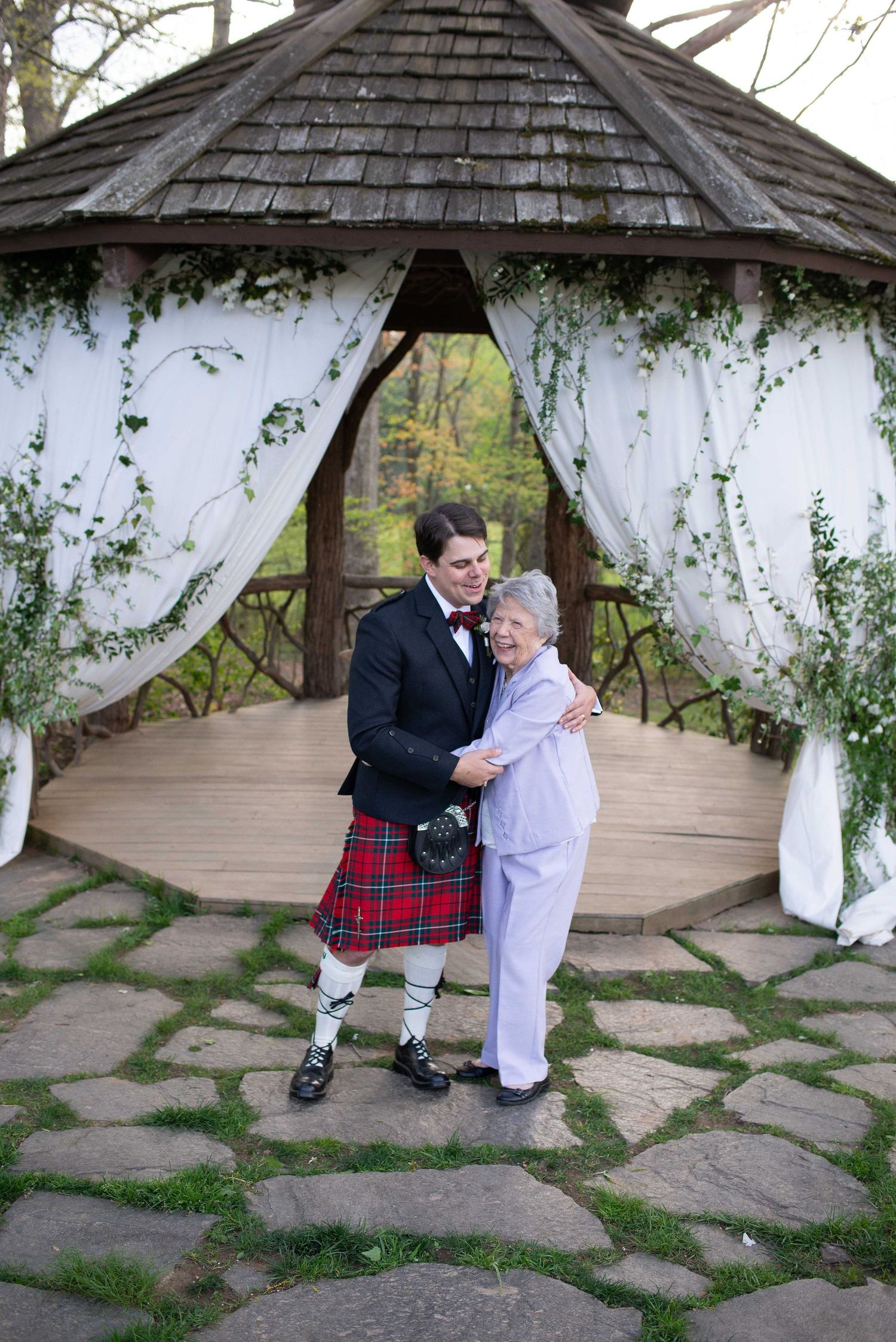 Couple embracing under decorated gazebo, man in Scottish kilt and tuxedo jacket, woman in white pantsuit, outdoor wedding.