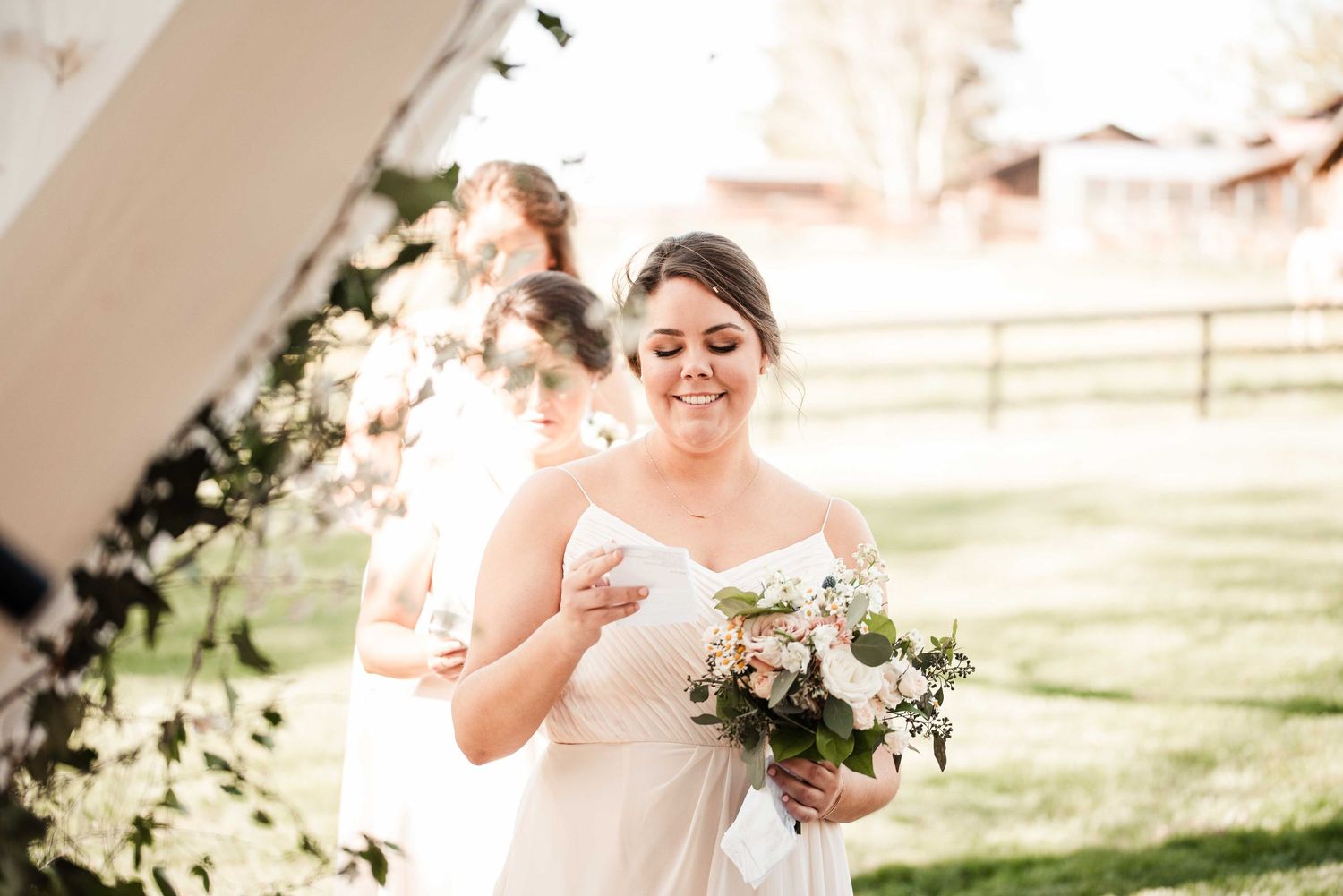Smiling bride in white gown holding a blush floral bouquet outdoors at a rustic barn wedding venue.