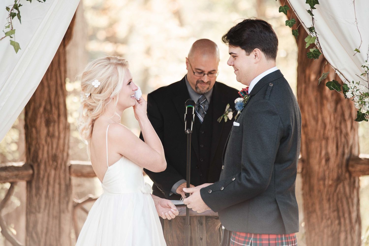 Bride and groom exchange vows outdoors under a floral arch, officiated by a man in a suit at a rustic wedding.