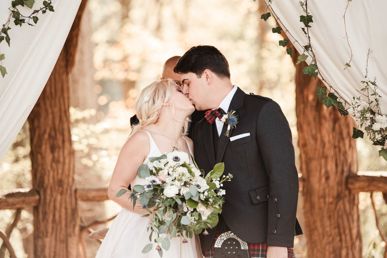 Bride and groom share first kiss at outdoor woodland wedding ceremony with white draping and greenery backdrop.