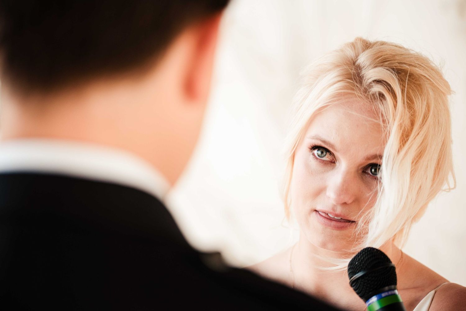 Blonde woman holding a microphone listens attentively to a man in a suit at a wedding ceremony.
