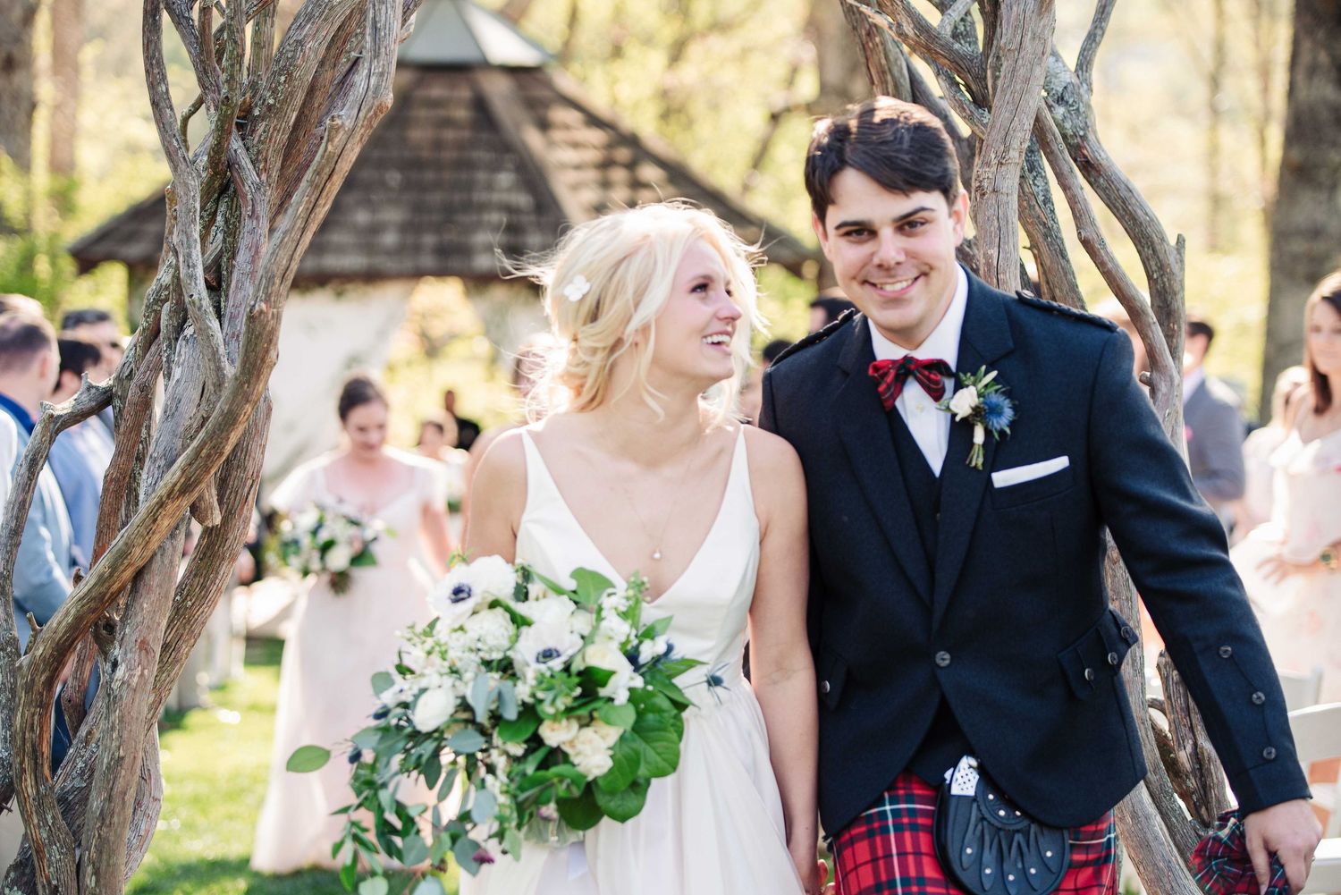 Smiling bride and groom in Scottish kilt walking down aisle after outdoor wedding ceremony.