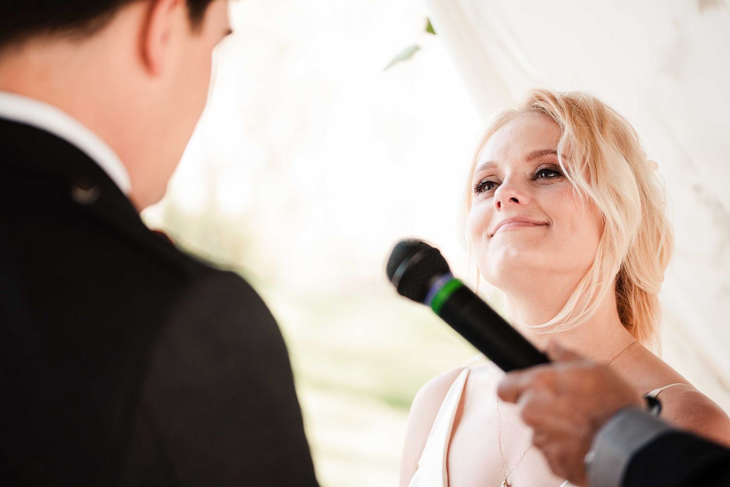 Blonde bride smiling emotionally while reciting wedding vows into a microphone during an outdoor ceremony.