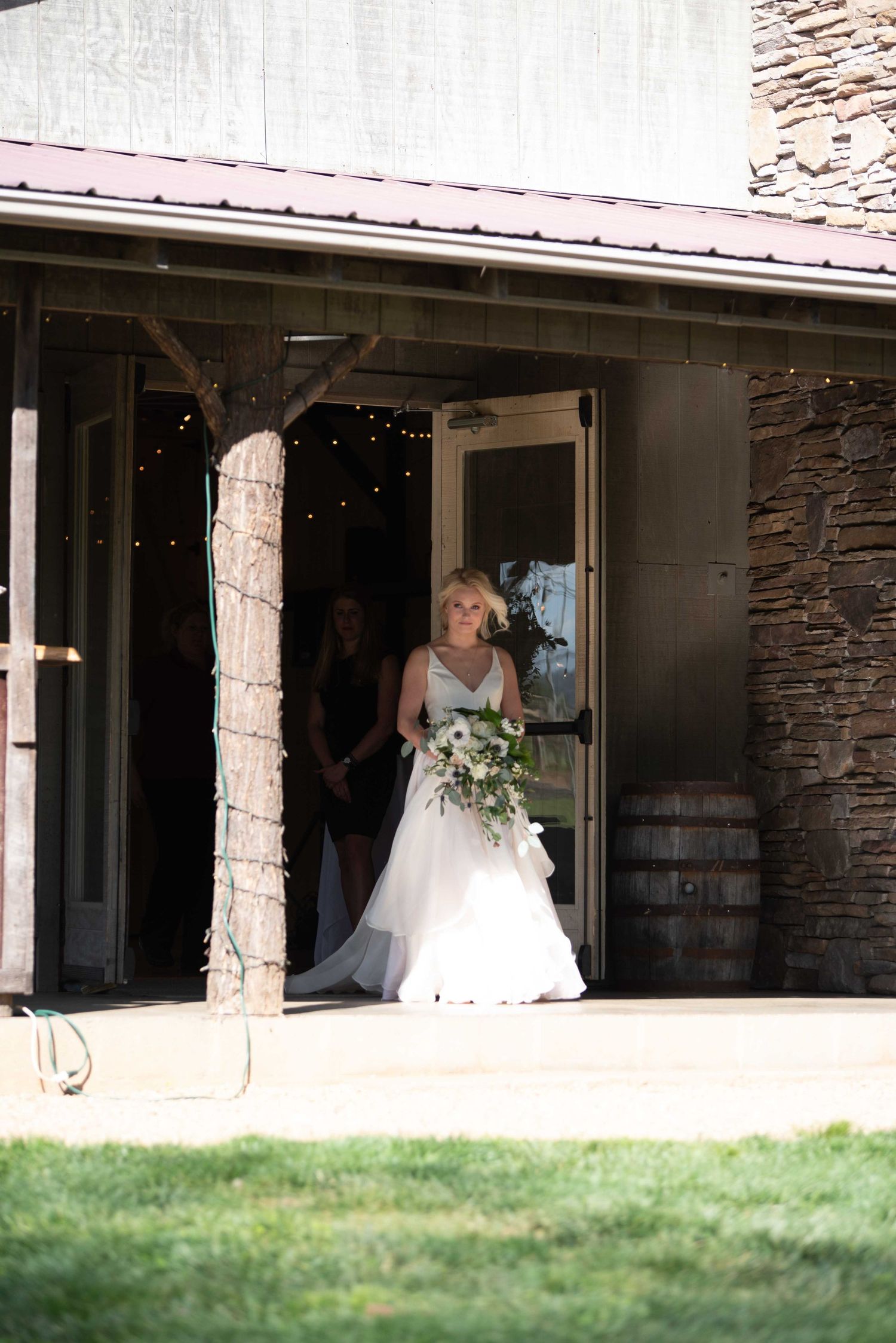 Bride in white gown holding bouquet exits barn venue doors with bridesmaid, string lights glowing inside.
