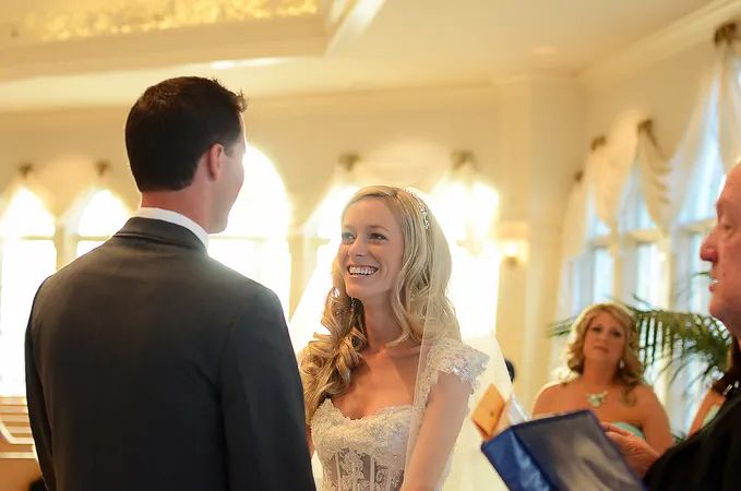 Radiant blonde bride laughing with groom during elegant indoor wedding ceremony in ornate venue.