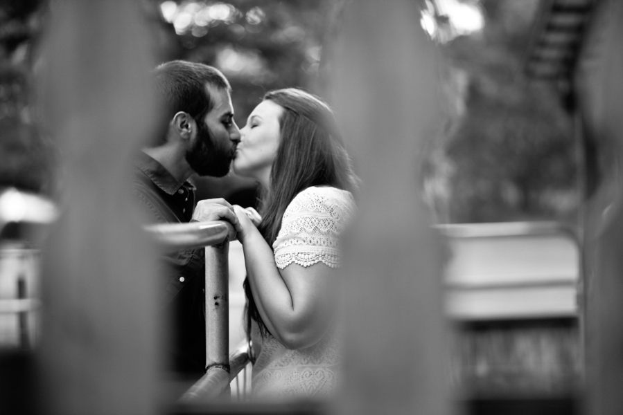 Black and white photo of a couple sharing a romantic kiss over a railing, captured through a wooden fence.