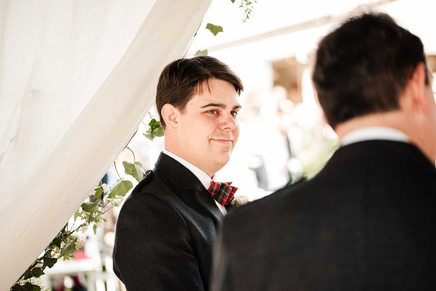 Groom in black tuxedo with floral bow tie smiling at wedding ceremony under white tent with greenery.
