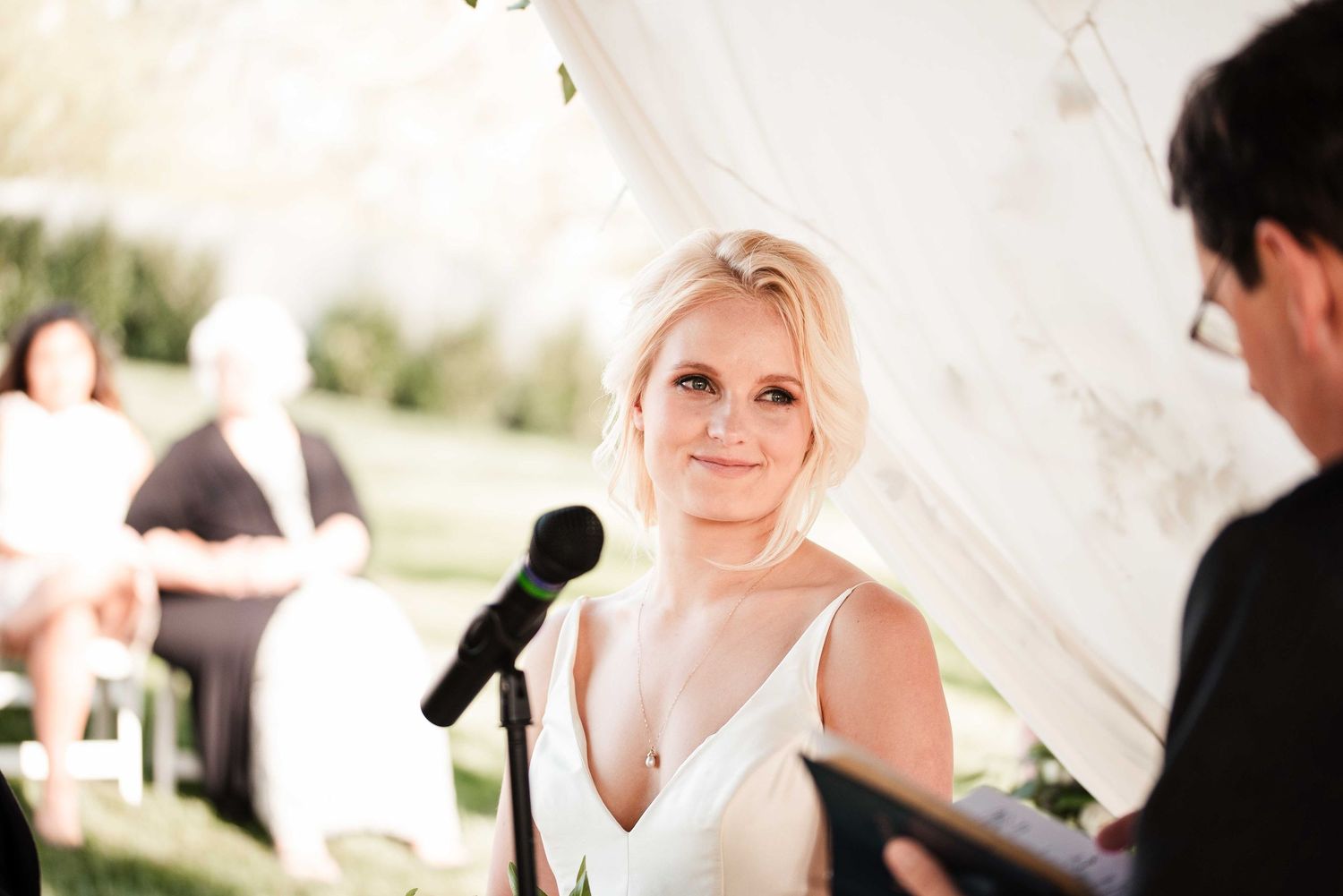 Bride smiling warmly at wedding ceremony under white tent, wearing elegant V-neck gown near microphone.