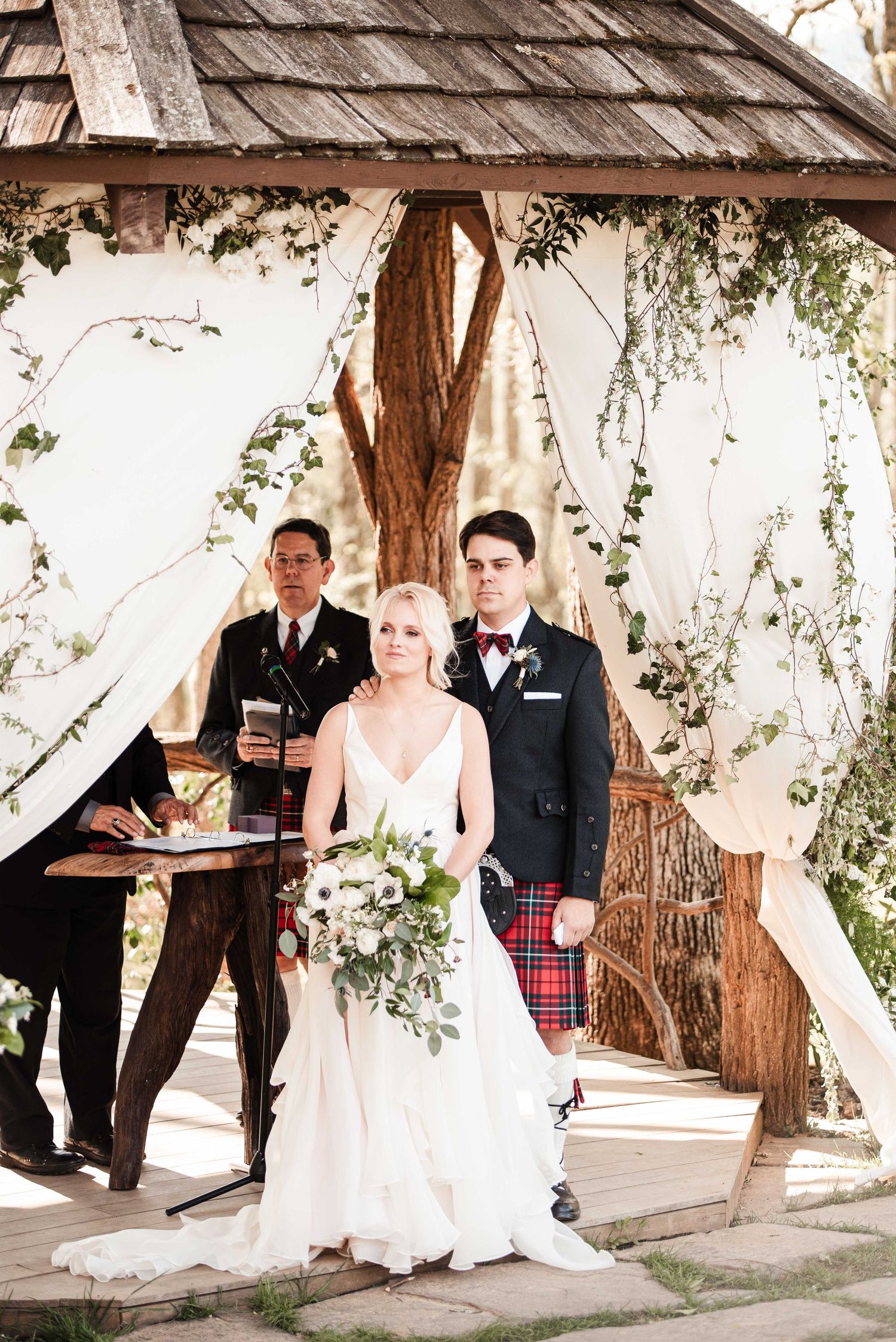 Bride in white gown holding bouquet stands with groom in kilt at rustic outdoor wedding ceremony under wooden gazebo.