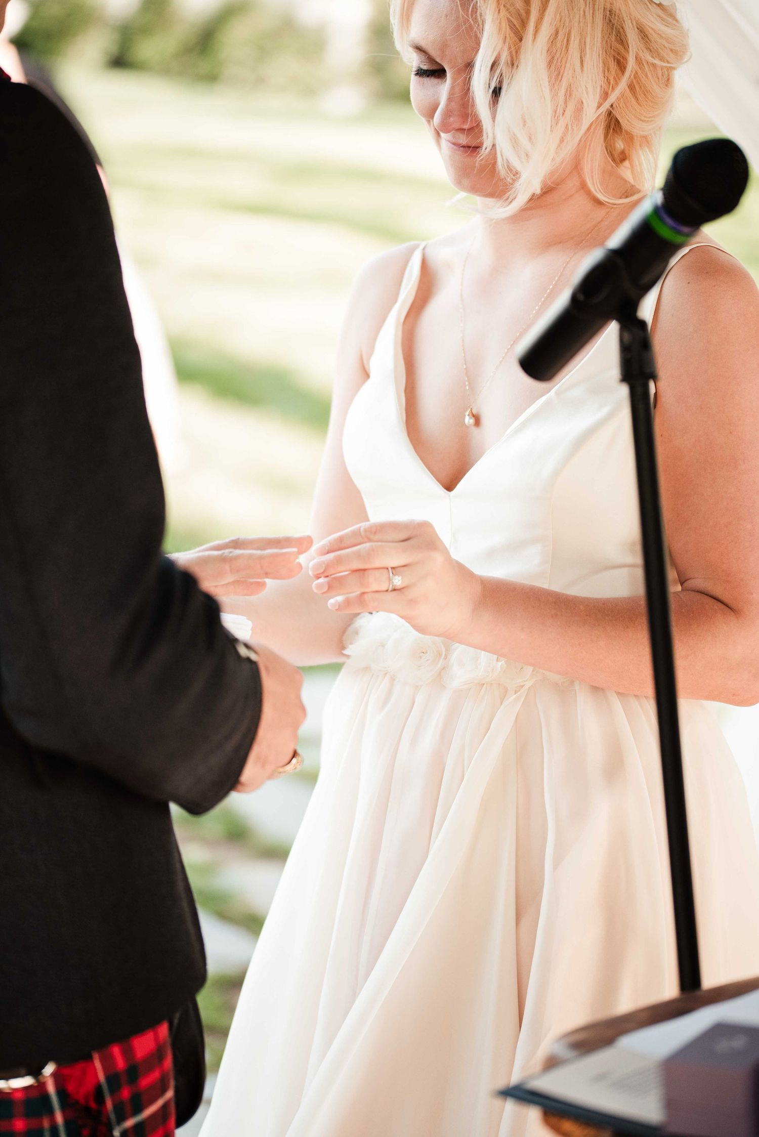 Smiling blonde bride in white gown placing wedding ring on groom's finger during outdoor ceremony.