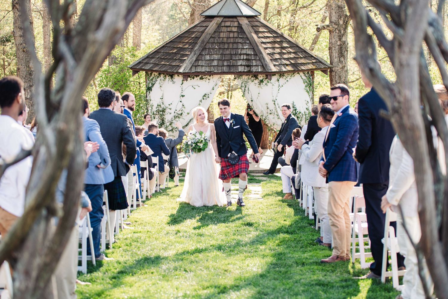 Bride and groom walk down outdoor aisle under gazebo surrounded by guests at a sunny garden wedding ceremony.