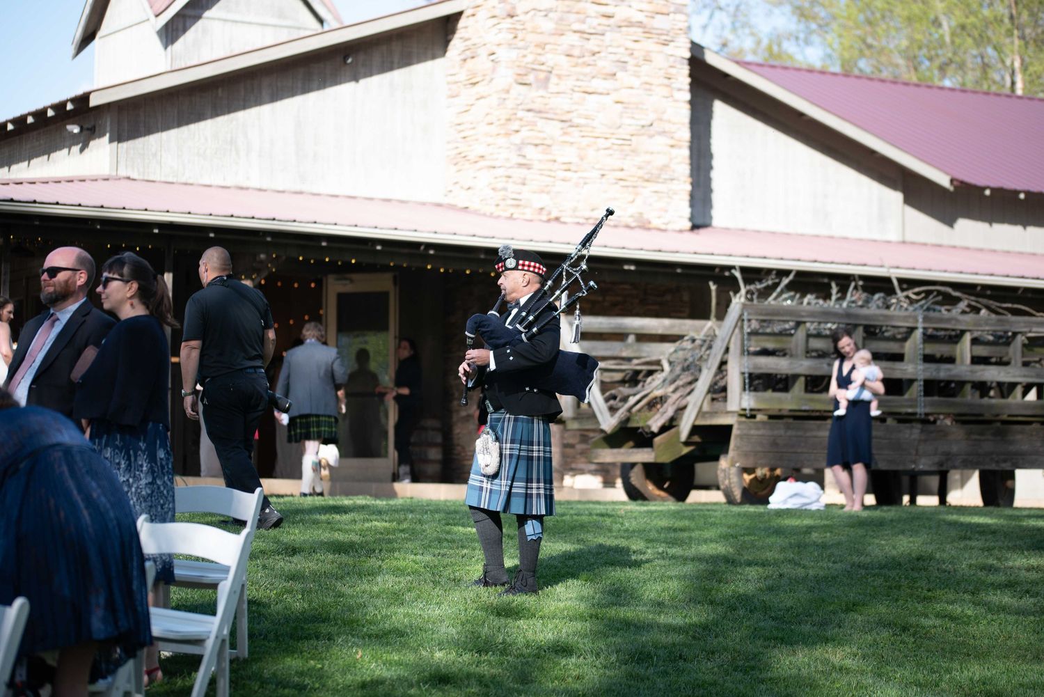 A bagpiper in traditional Scottish kilt performs outdoors at a wedding venue with guests gathered near a rustic barn.