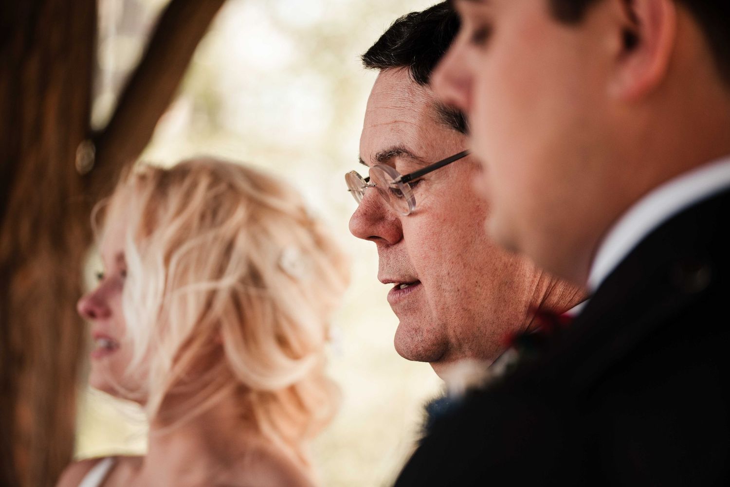 Close-up of wedding guests, including a blonde woman and a man wearing glasses, during an outdoor ceremony.