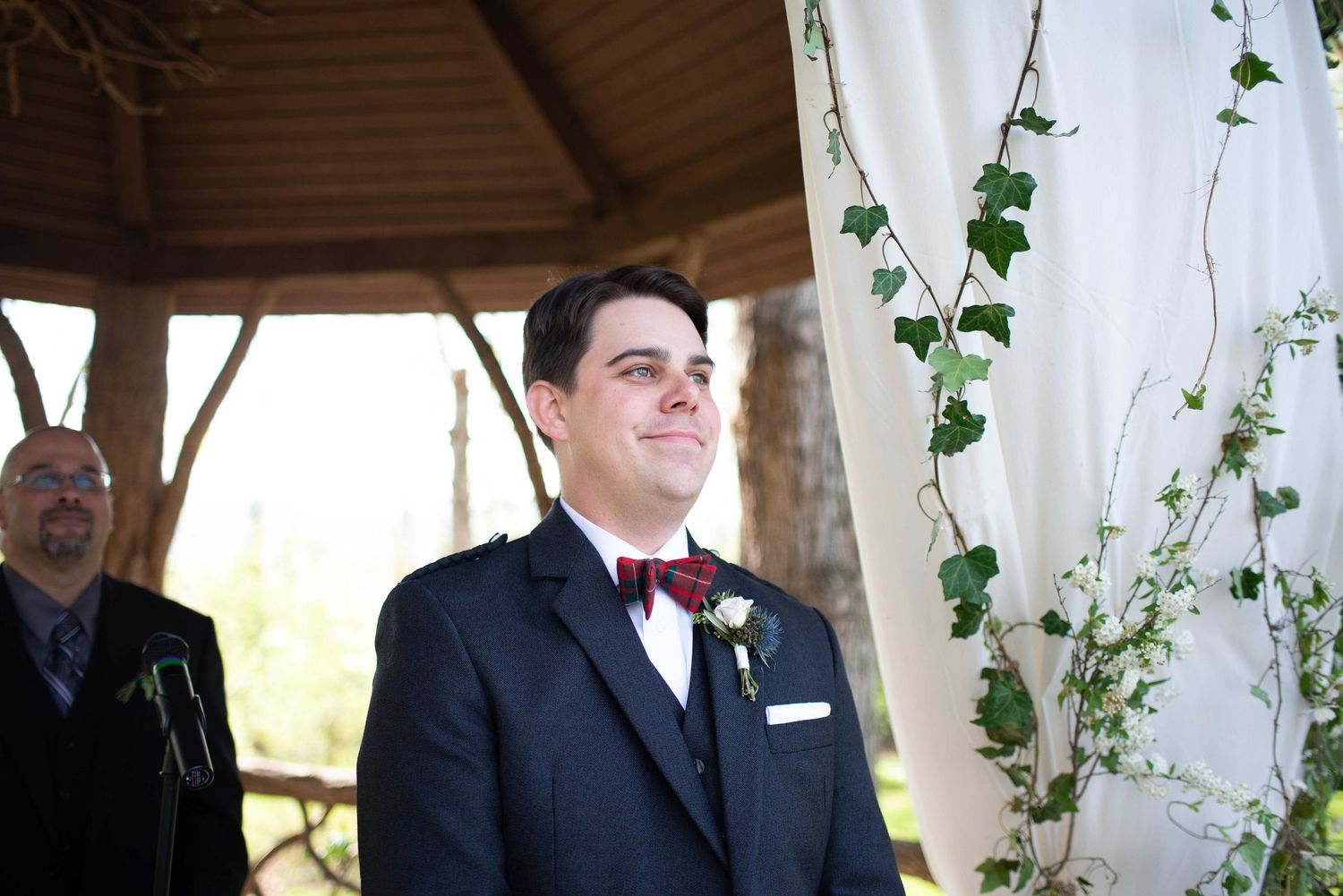 Groom in navy suit with burgundy boutonniere smiling at altar decorated with ivy during outdoor wedding ceremony.