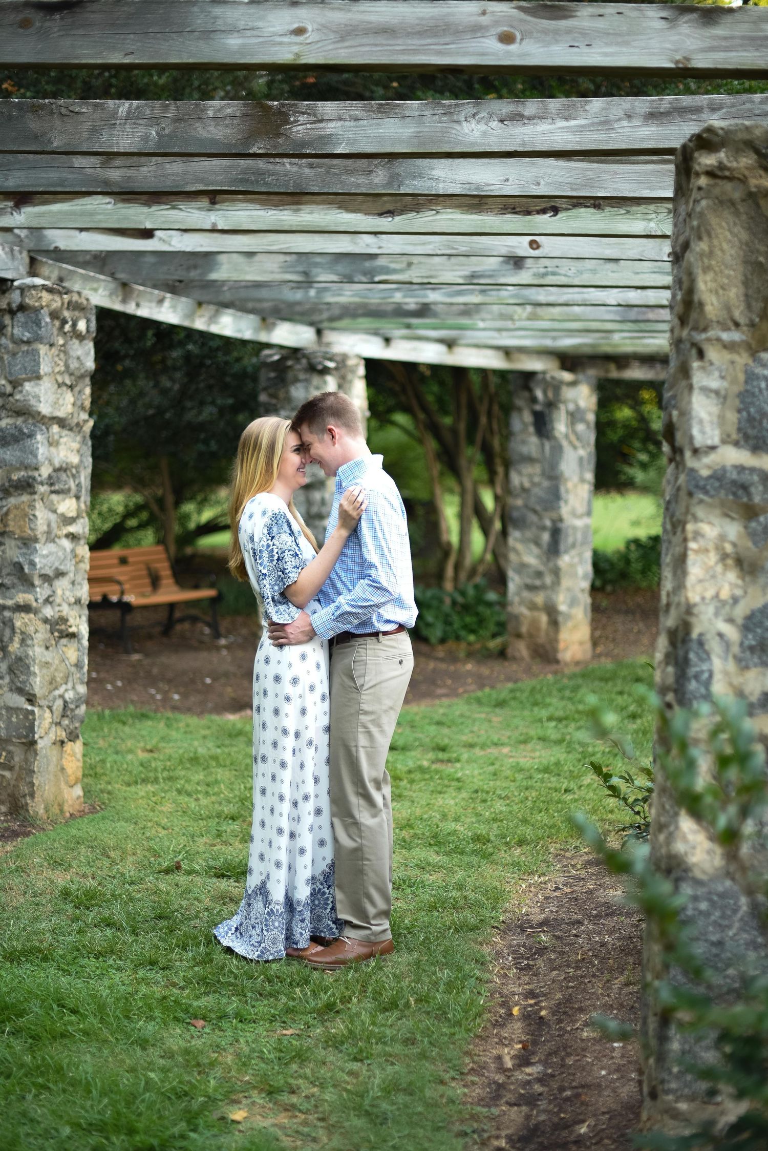 Couple sharing an intimate moment under a rustic wooden pergola with stone columns in a garden.