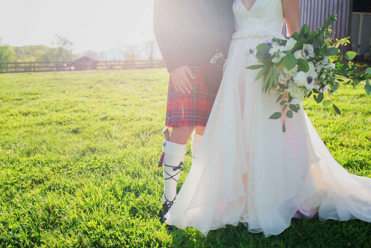 A groom in a traditional Scottish kilt stands beside his bride holding a white floral bouquet outdoors.