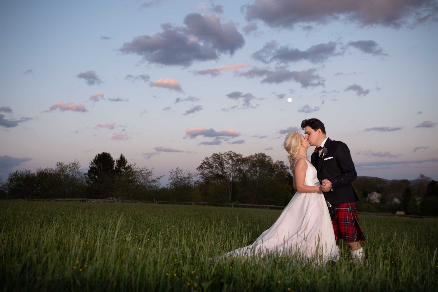 Bride and groom share a romantic kiss in a green field at sunset, groom wearing a traditional red tartan kilt.