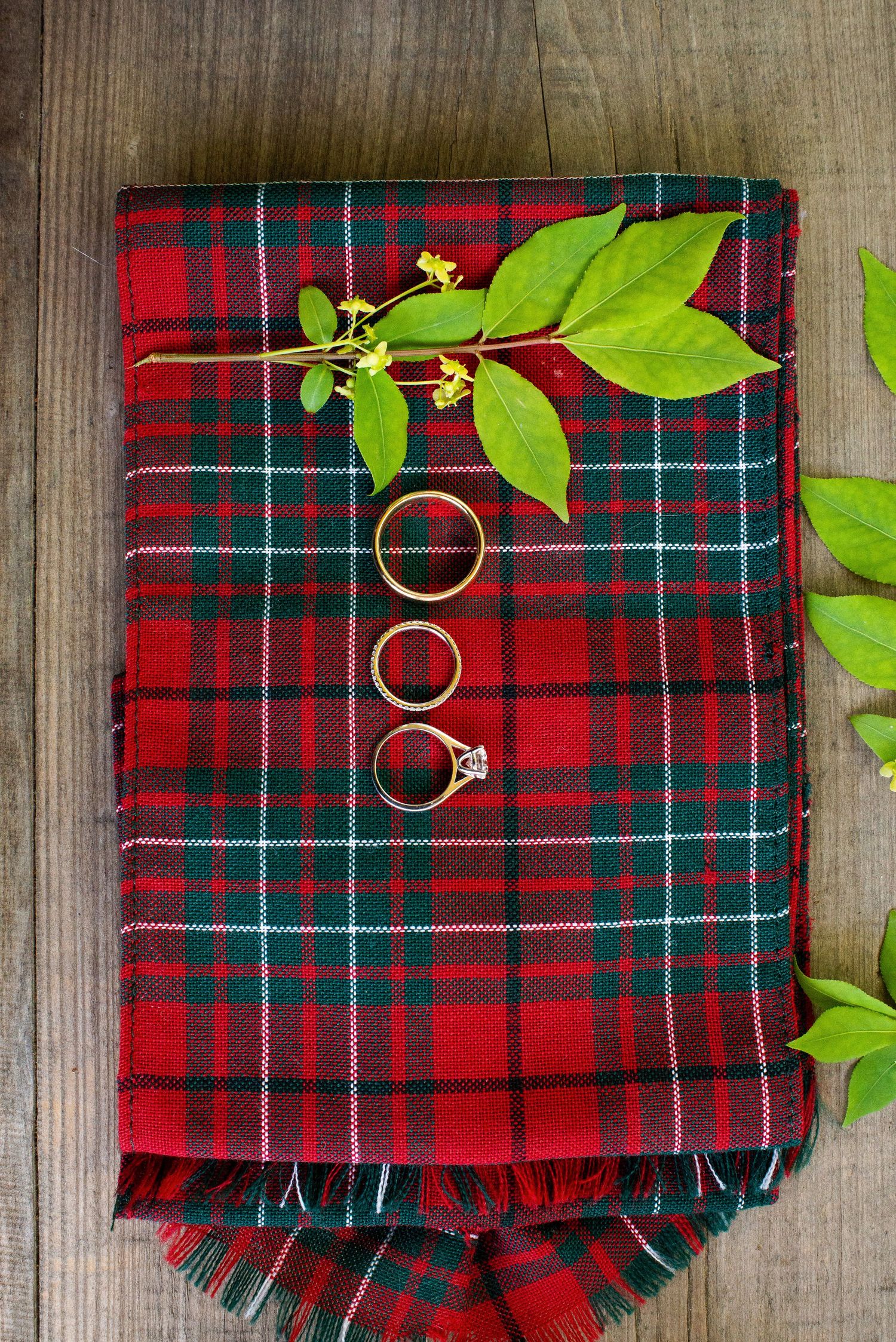 Gold wedding rings and diamond engagement ring displayed on a folded red tartan plaid fabric with green leaves.