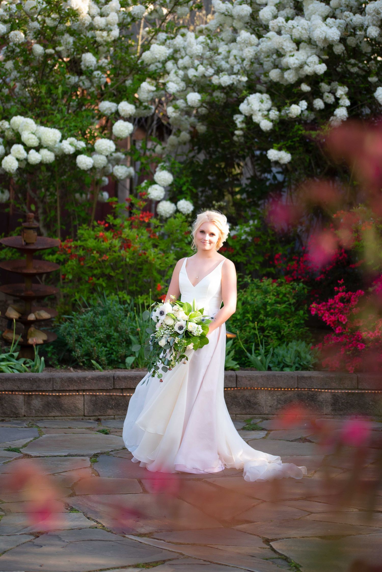 Bride in white gown holding bouquet stands on stone patio surrounded by lush white and pink flowering garden.