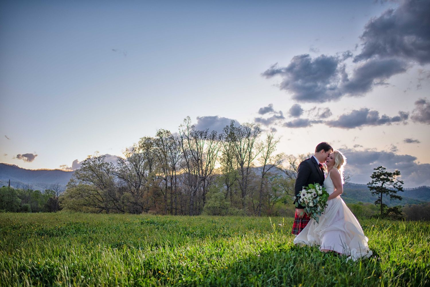 Bride and groom share a romantic kiss in a lush green field with mountain views and a beautiful sunset sky.