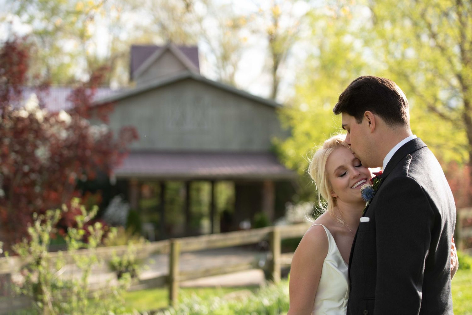 A bride and groom share a tender embrace outdoors near a rustic barn surrounded by lush green trees on their wedding day.