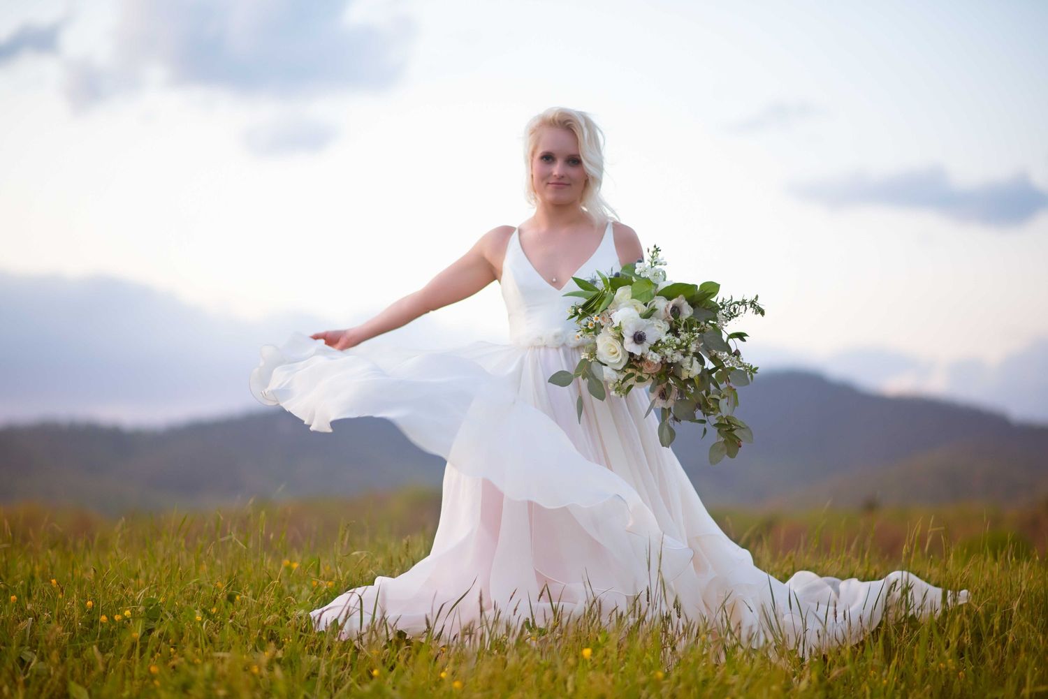Bride in flowing white gown holding bouquet in mountain meadow.
